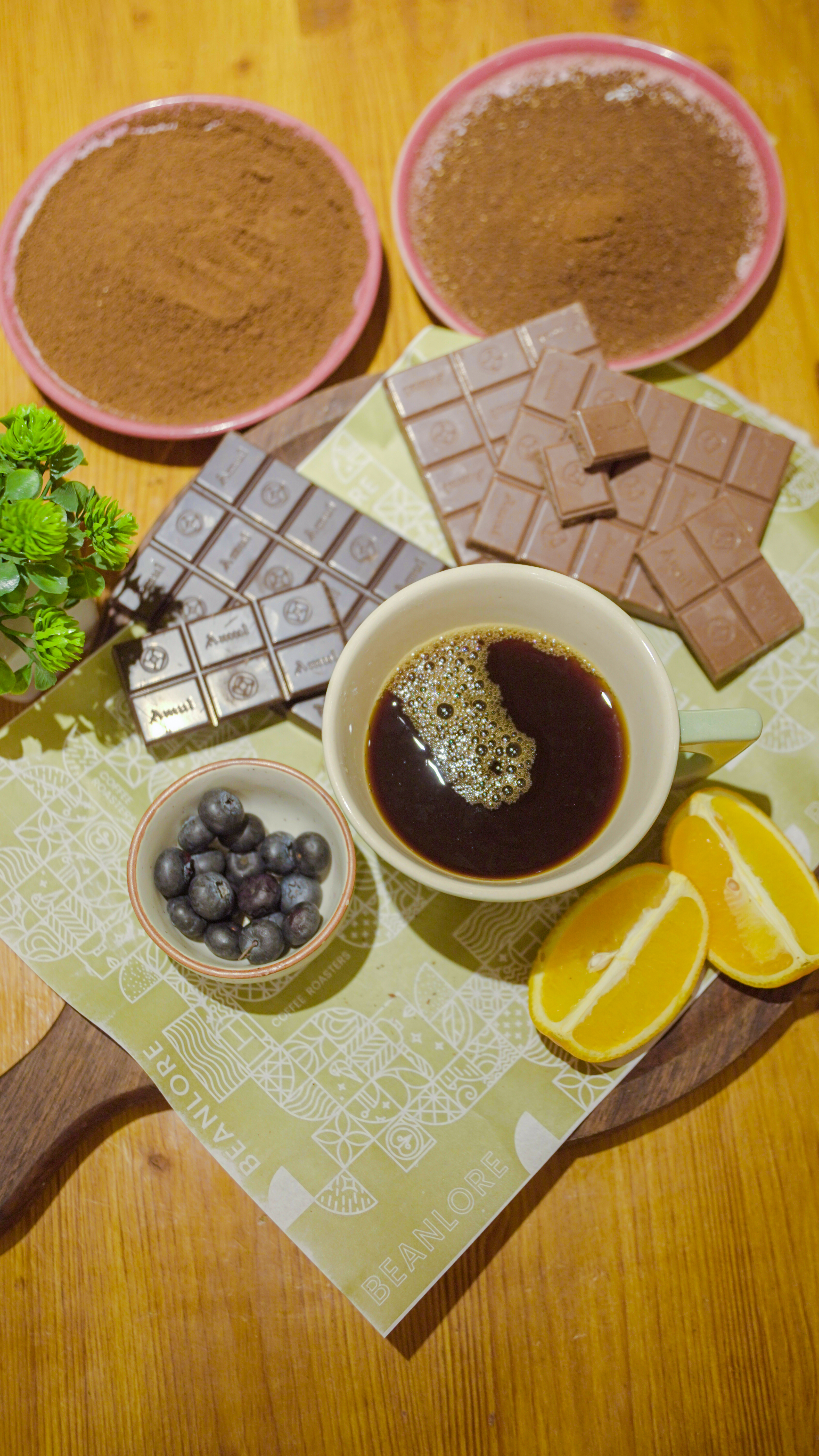 Ingredients including coffee powder, chocolate, blueberries and orange slices arranged around a cup of brewed coffee on a table.
